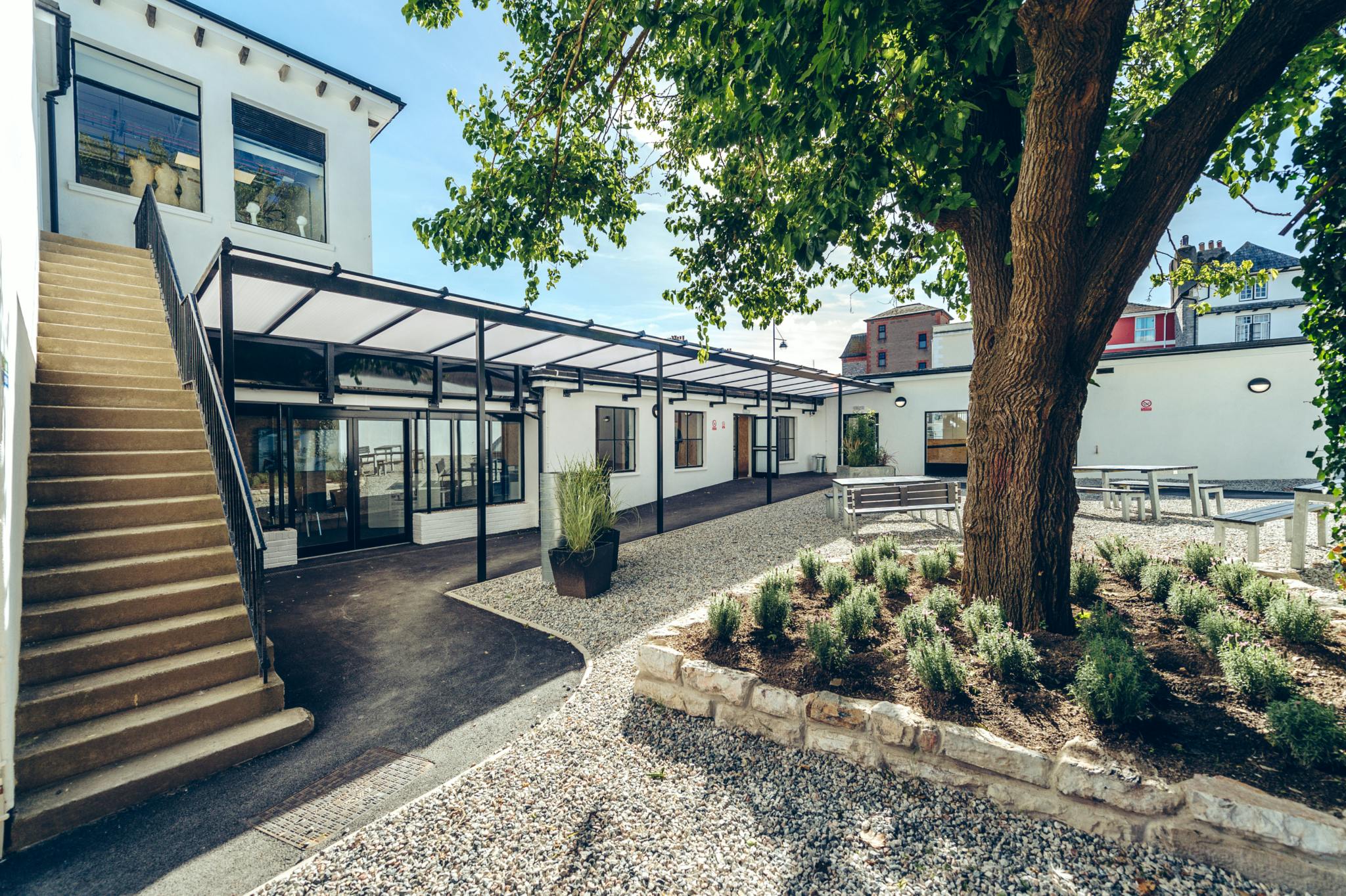 Palace Court’s outdoor courtyard basks in the summer sun. A golden-brown staircase to the left leads to a second-floor entranceway, whilst ground-level a large white mulberry tree is surrounded by pale grey gravel and white walls with black framing.