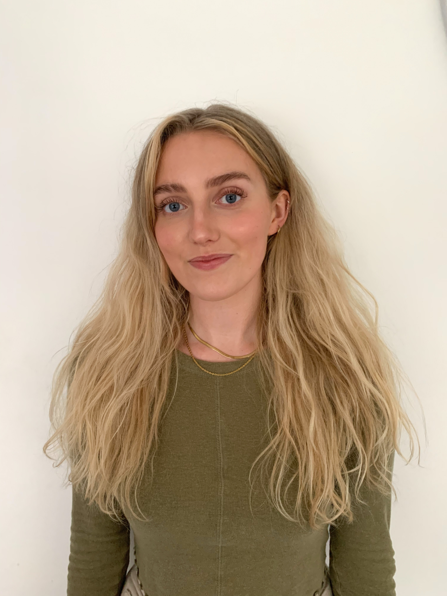 Image shows a woman with long blonde hair and blue eyes wearing a long sleeved olive green top with a small smile on her face looking straight at the camera in front of a white background