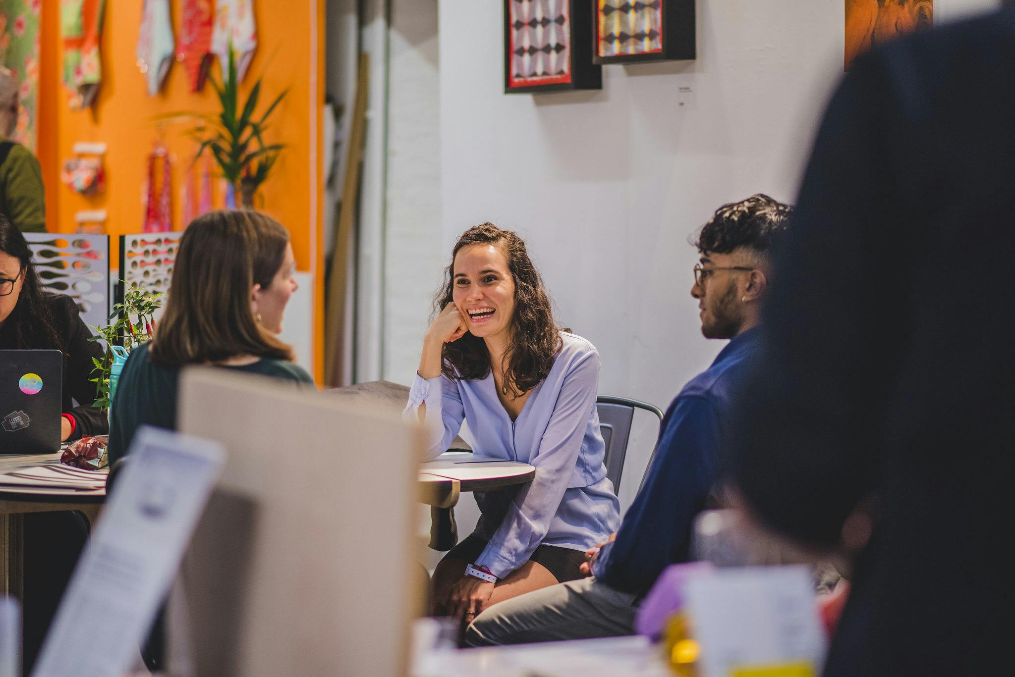 A group of industry visitors gather around a table, surrounded by student work and a vibrant orange wall.