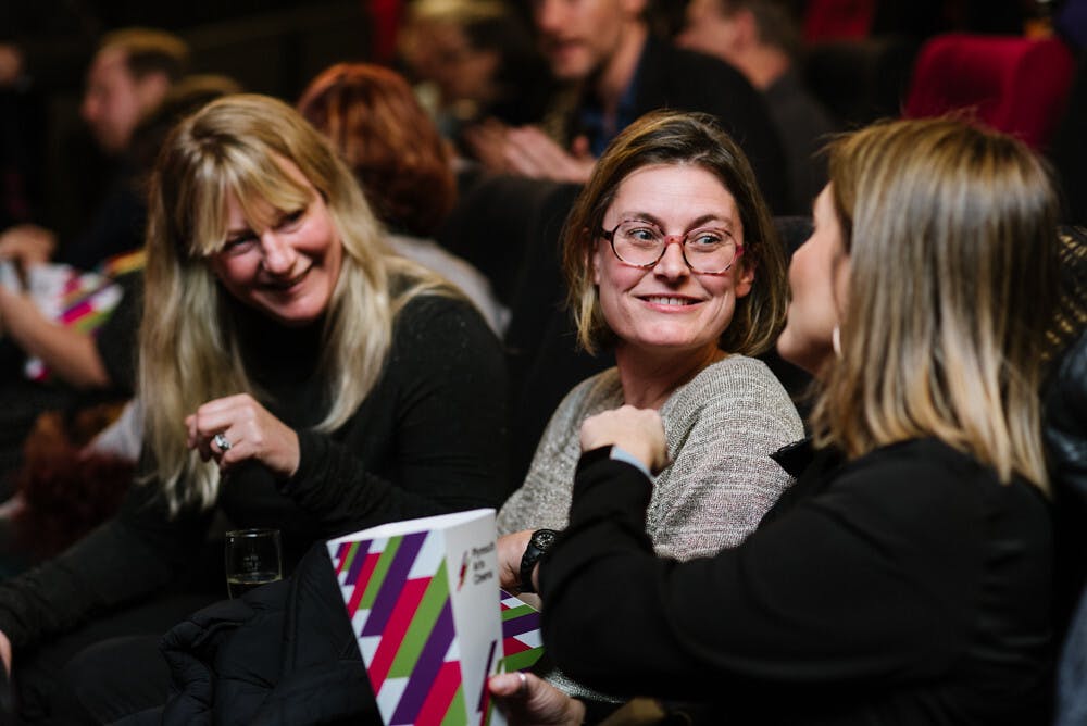 Three women eat popcorn and chat to eachother in their cinema seats. They wear black and grey jumpers.