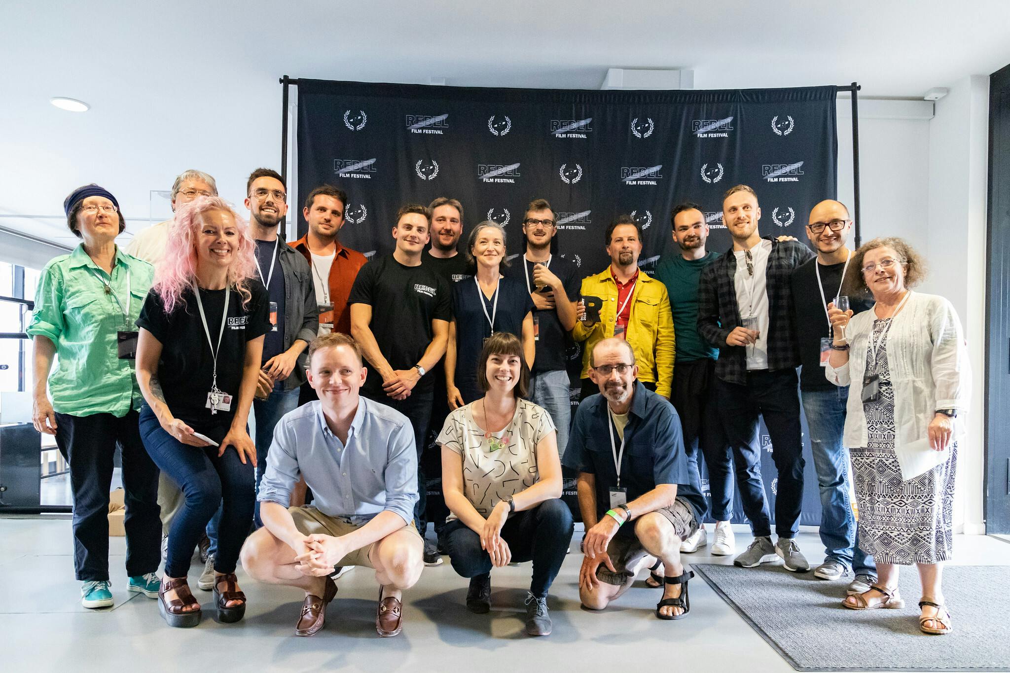 A group photo in the college reception space flooded with natural light. 19 people stand in front of a black backdrop with white logos.