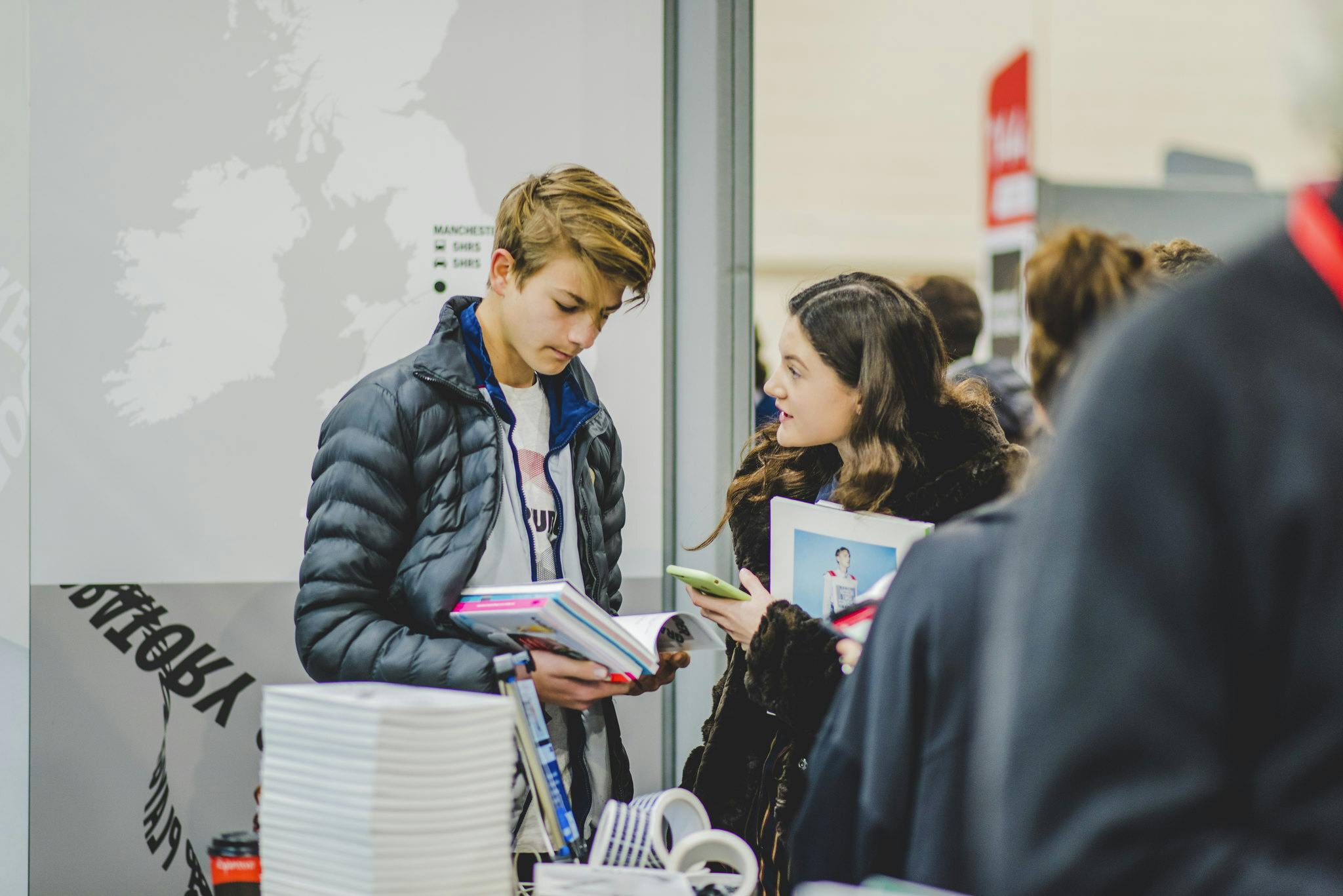 Two young people stand at a busy event looking at Plymouth College of Art prospectus and having a conversation with eachother.