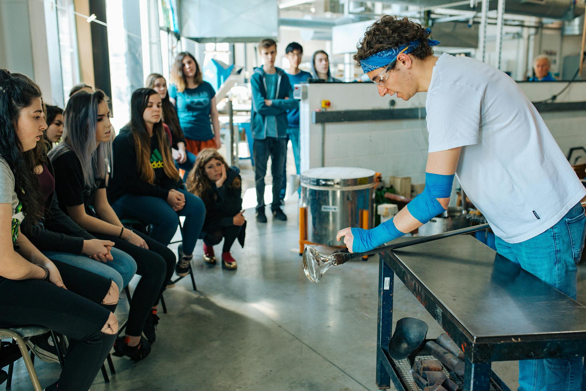 Plymouth College of Art glass alumnus Ben Lintell demonstrates glassblowing in our glass workshops, he is watched by students sat on chairs.