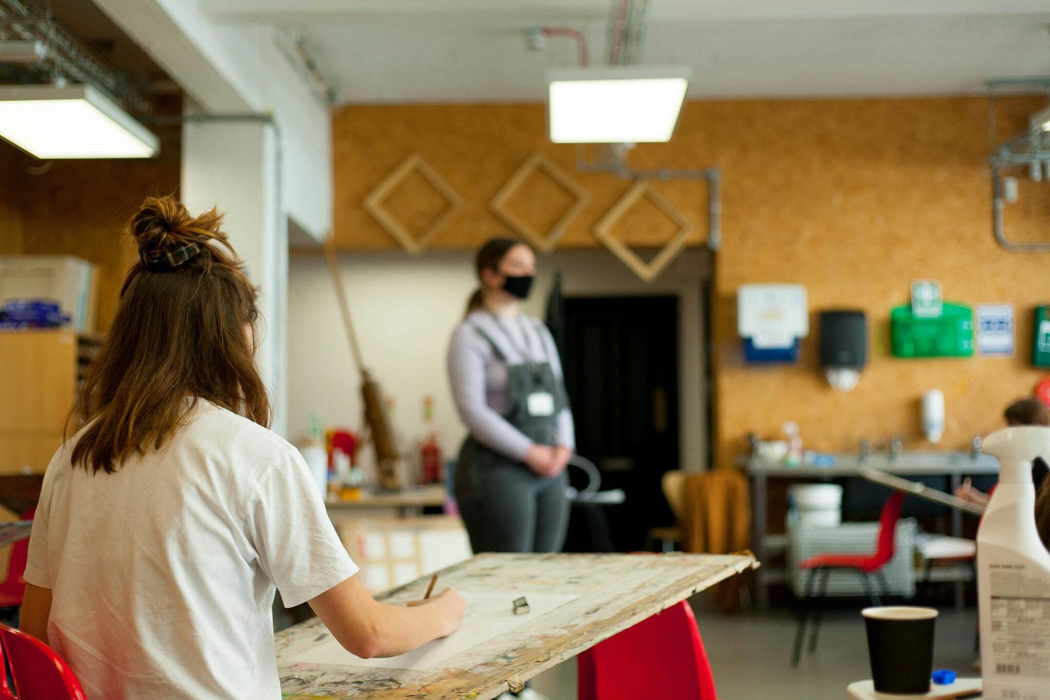 A woman in dungarees poses in the centre of the room for a life drawing session. The focus is on a young girl drawing on a large sheet of paper on an easel.