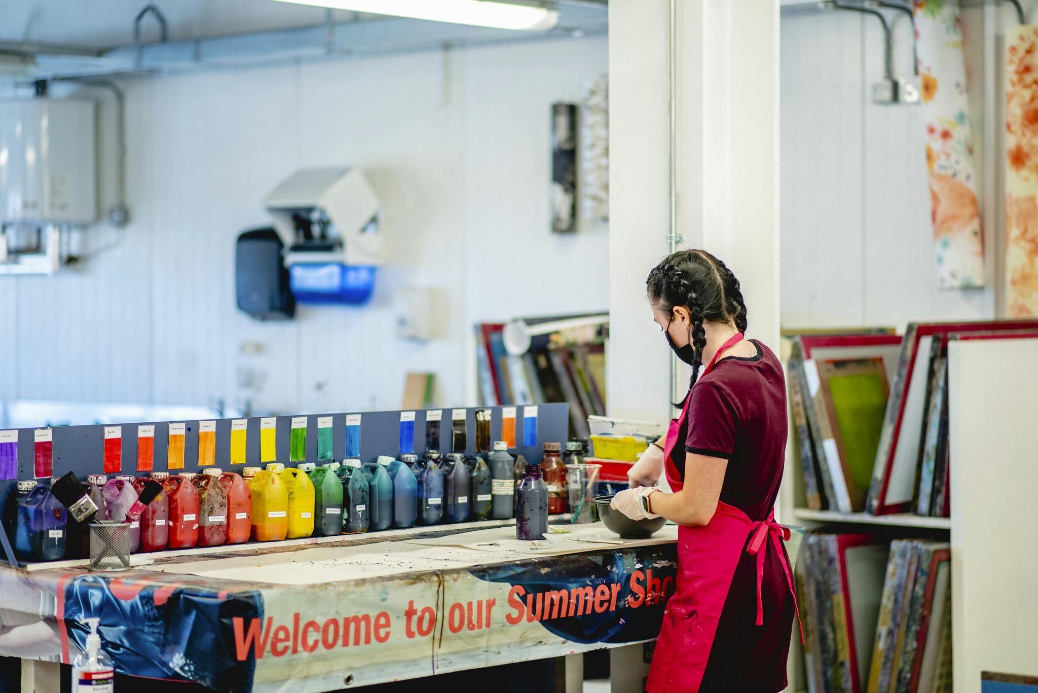 Pots of paint line up on a table in a rainbow of colours as a student, wearing a red apron, deposits some of the paint into a pot.