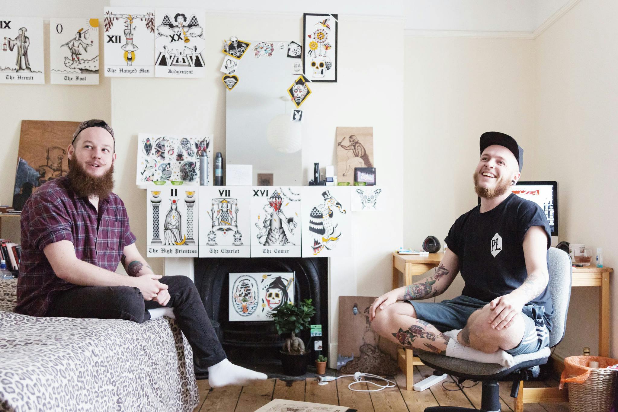 Two male students sit in a bedroom smiling and talking. The room is filled with natural light from the large window, tattoo style art adorns the walls and period features like the fireplace and oak floorboards give a homely feel.