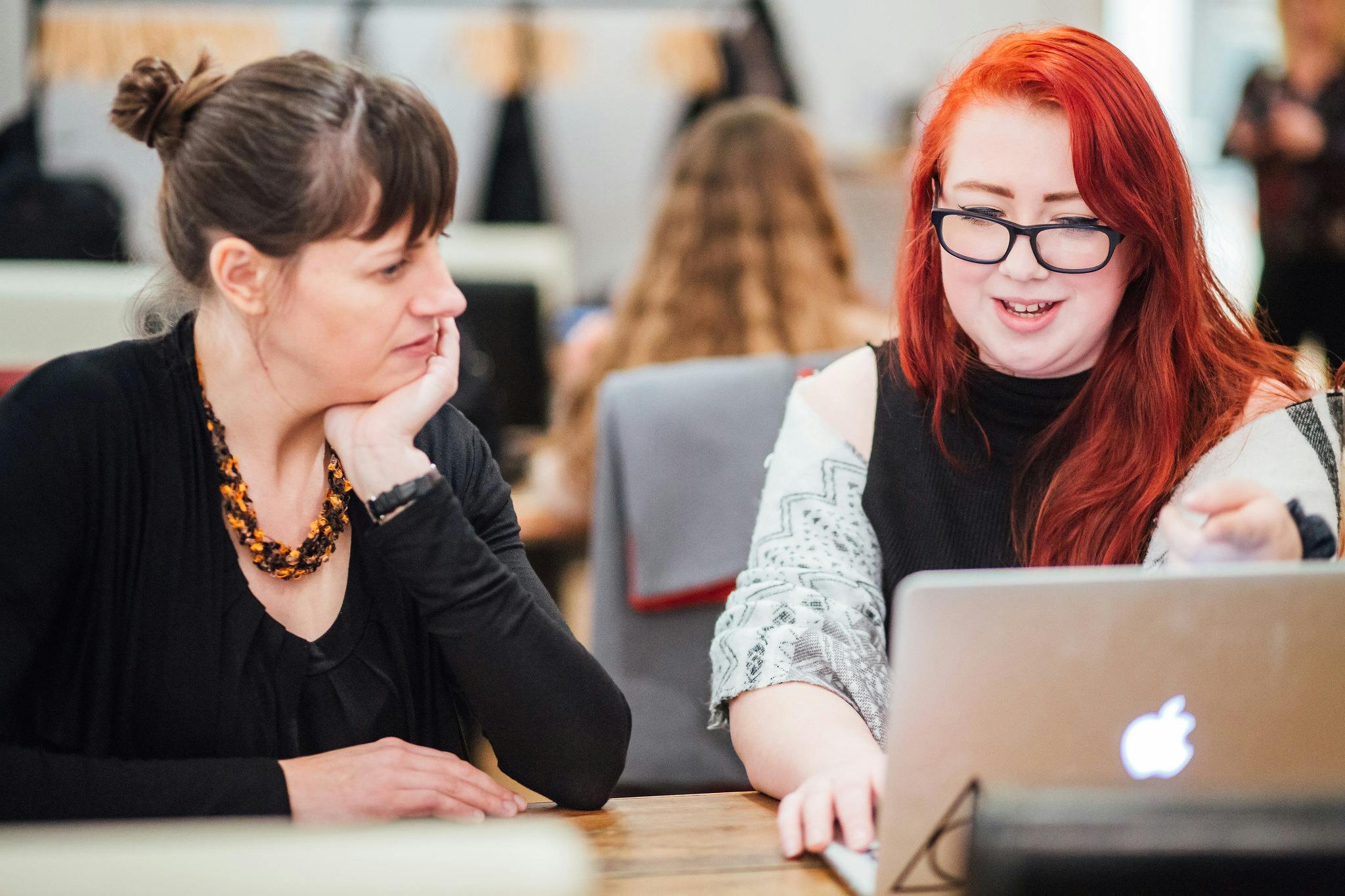 A girl with red hair and glasses smiles as she shows her portfolio on her laptop to the photographer sitting next to her.