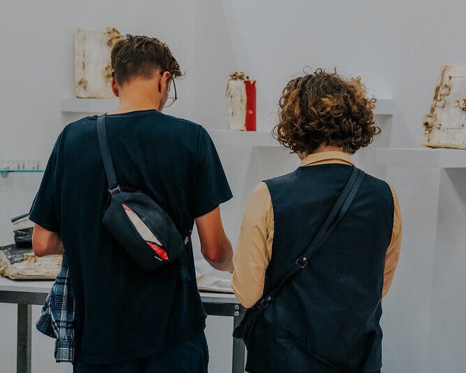 Two people wearing dark coloured outfits stand in a white walled gallery space with their backs to the camera