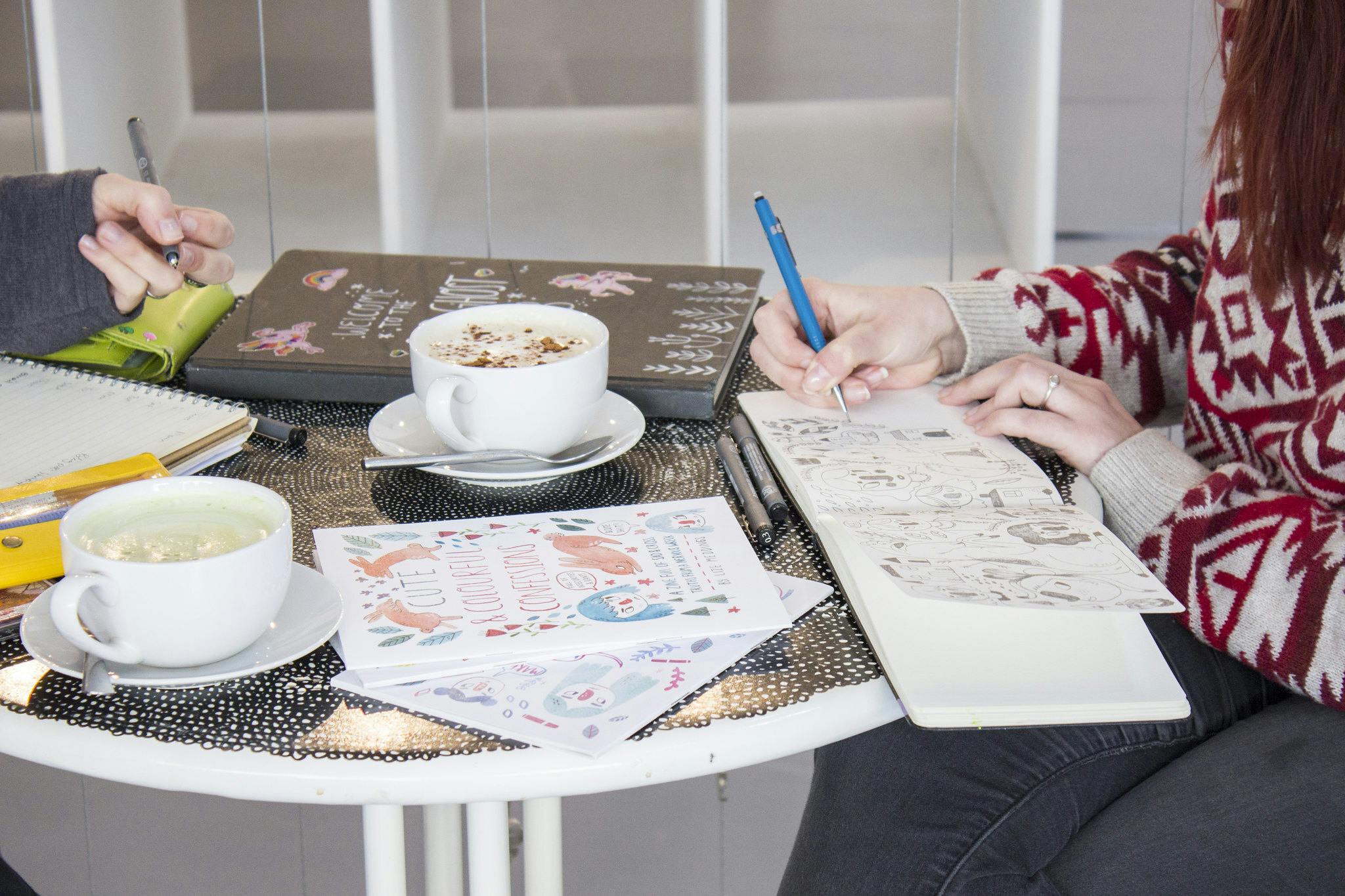 "A coffee table is crammed with sketchbooks and two cups of latte. Two young women sit at the table drawing in a sketchbook."