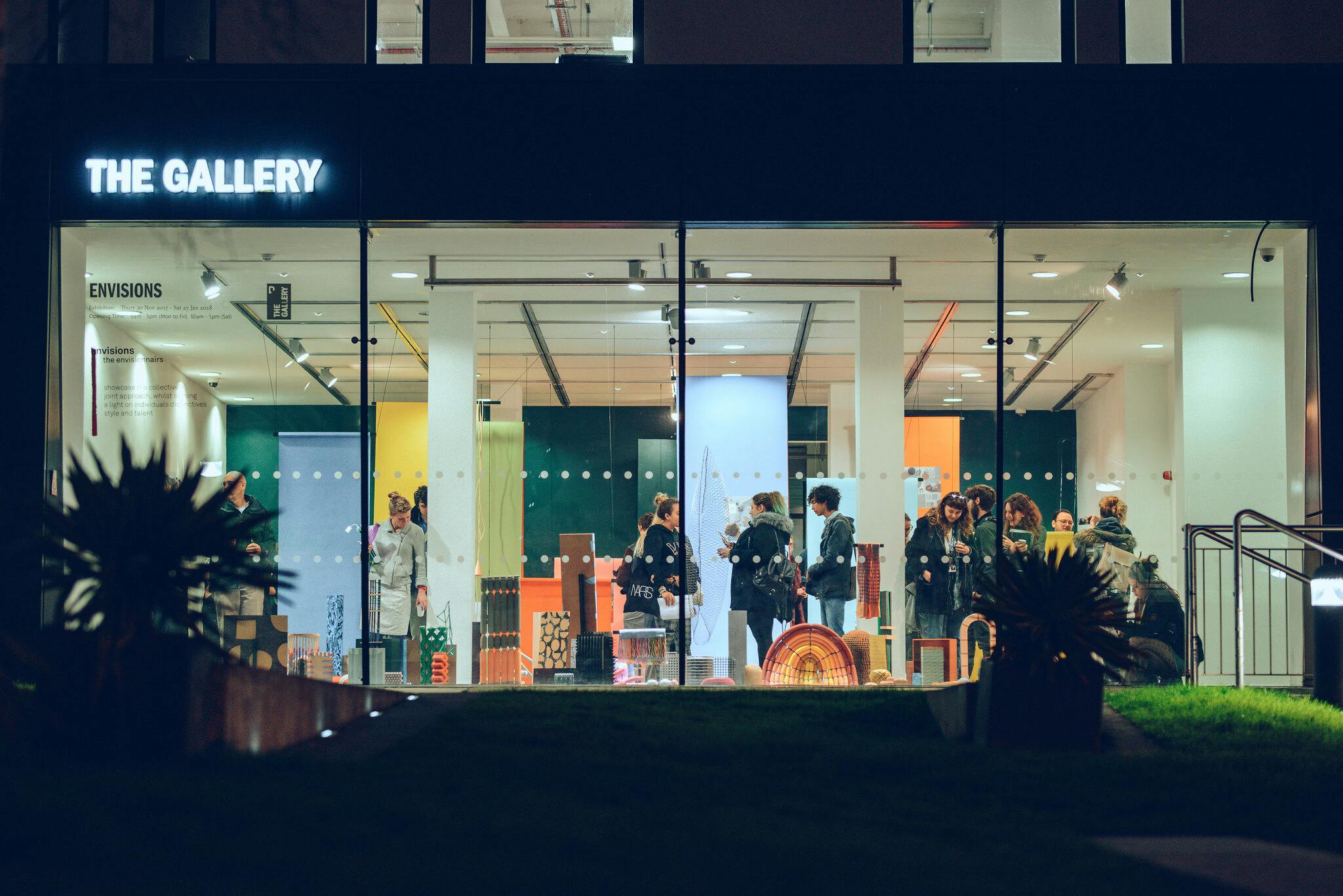 A lively exhibition opening is lit up in the dark of the evening, the floor to ceiling gallery windows show colourful sculptural pieces as guests chat and enjoy the space.