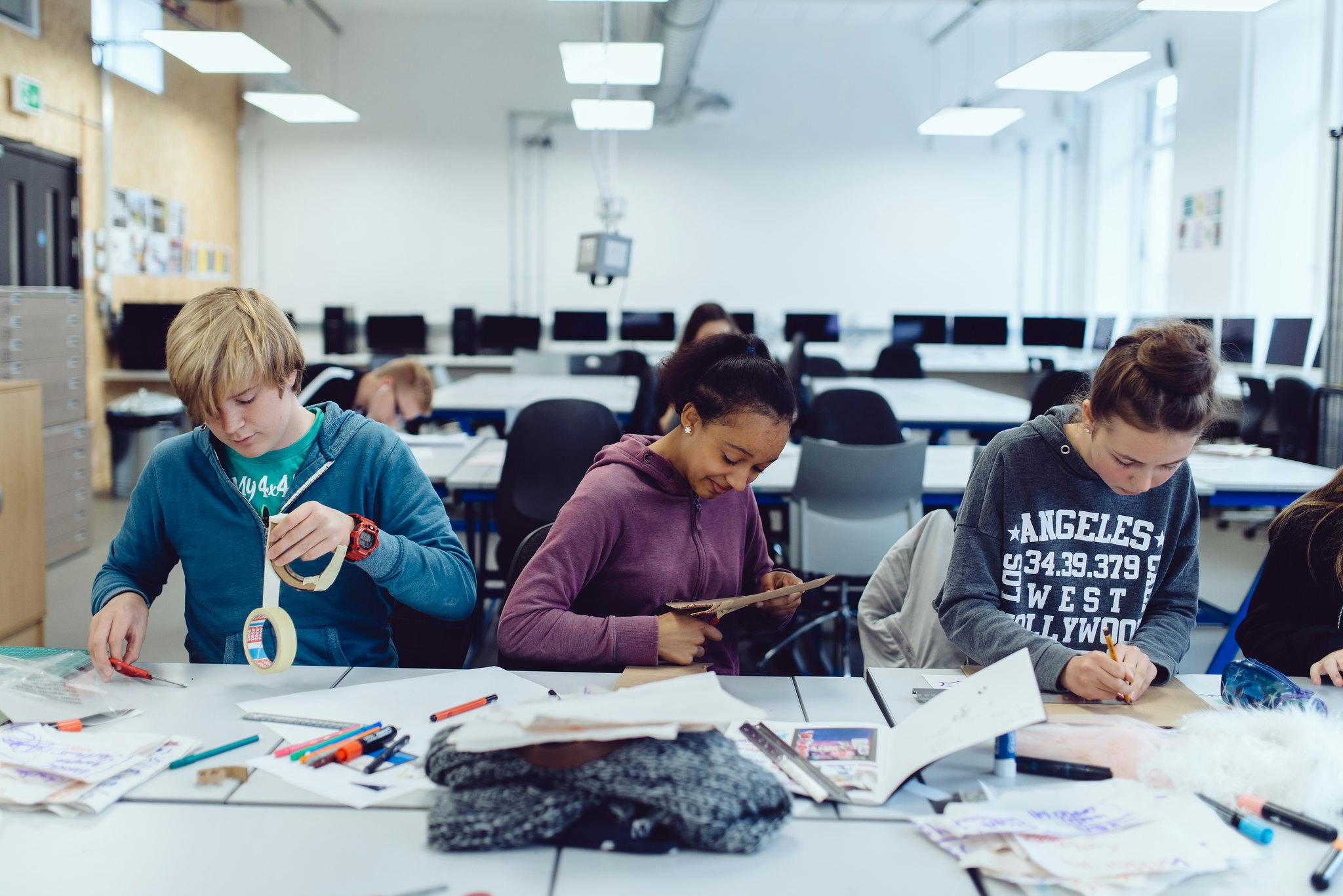 Three young people concentrate in a row at a table, cutting, sticking and taping their sketchbooks.