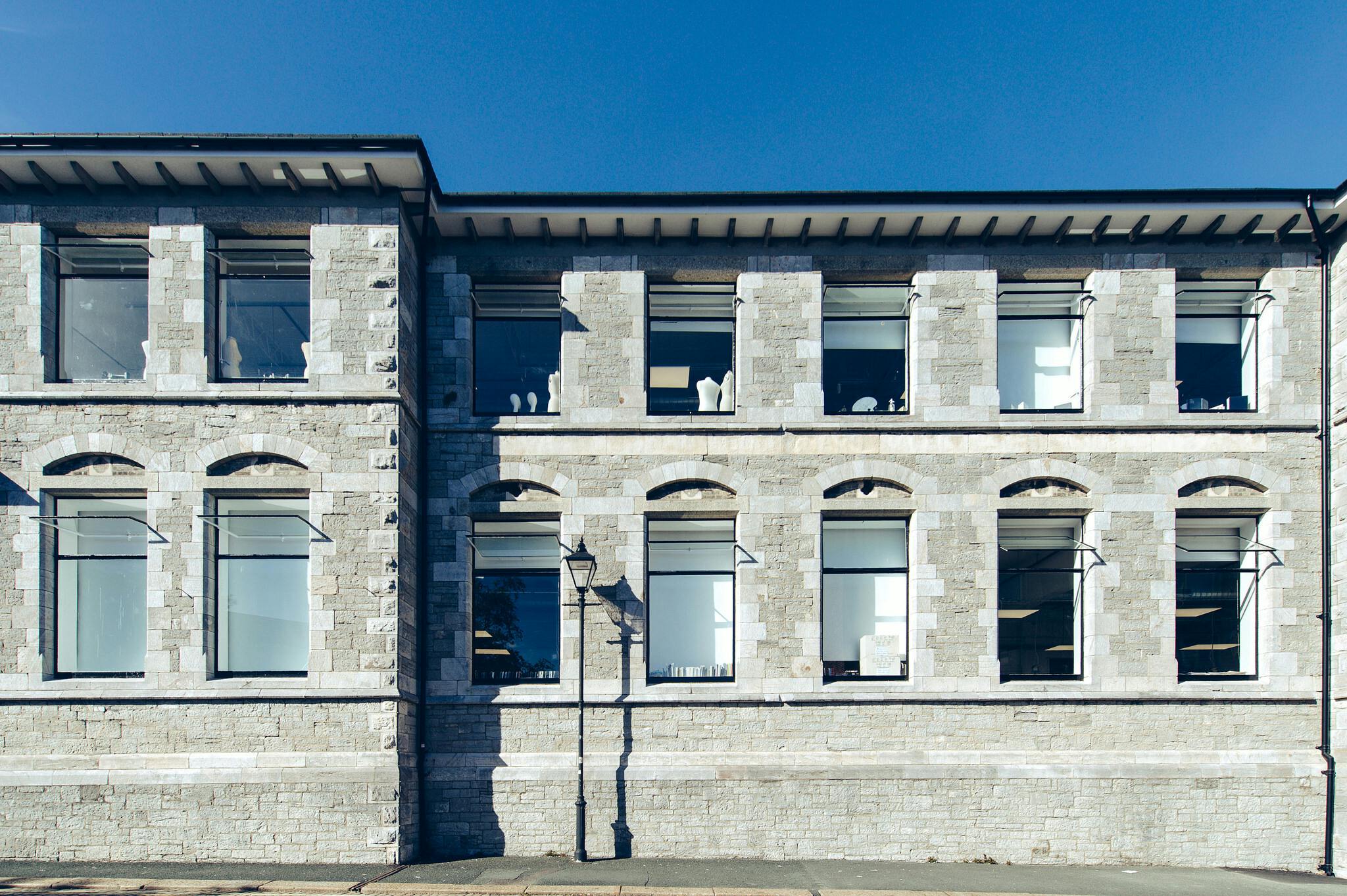 Palace Court's historic grey stone walls peer out against a bright blue sky. Two storeys of arched windows and an old fashioned lamppost can be seen.