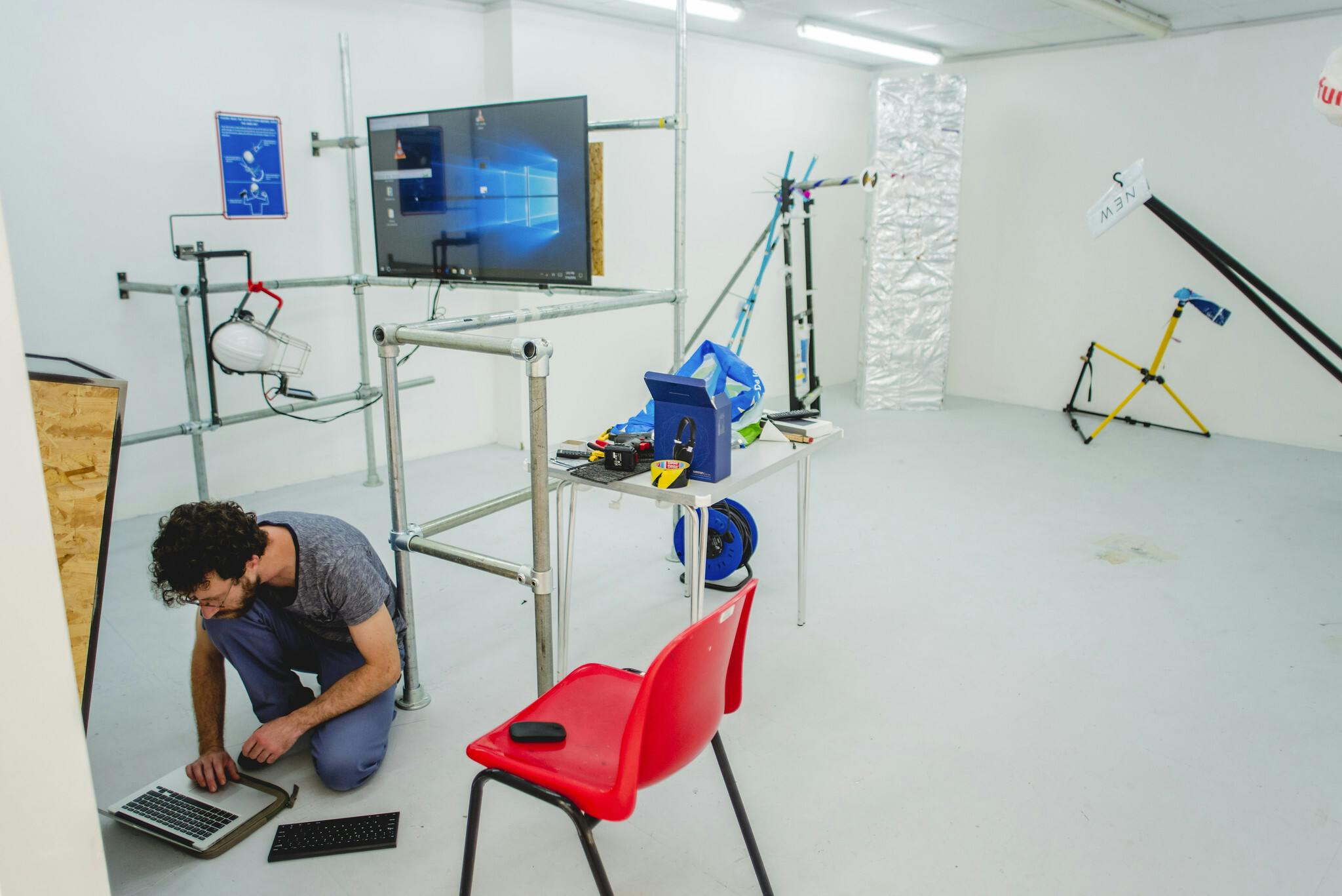 A male student kneels on the floor at his laptop, surrounded by carefully placed scaffolding, a wide-screen TV and a bright red chair.