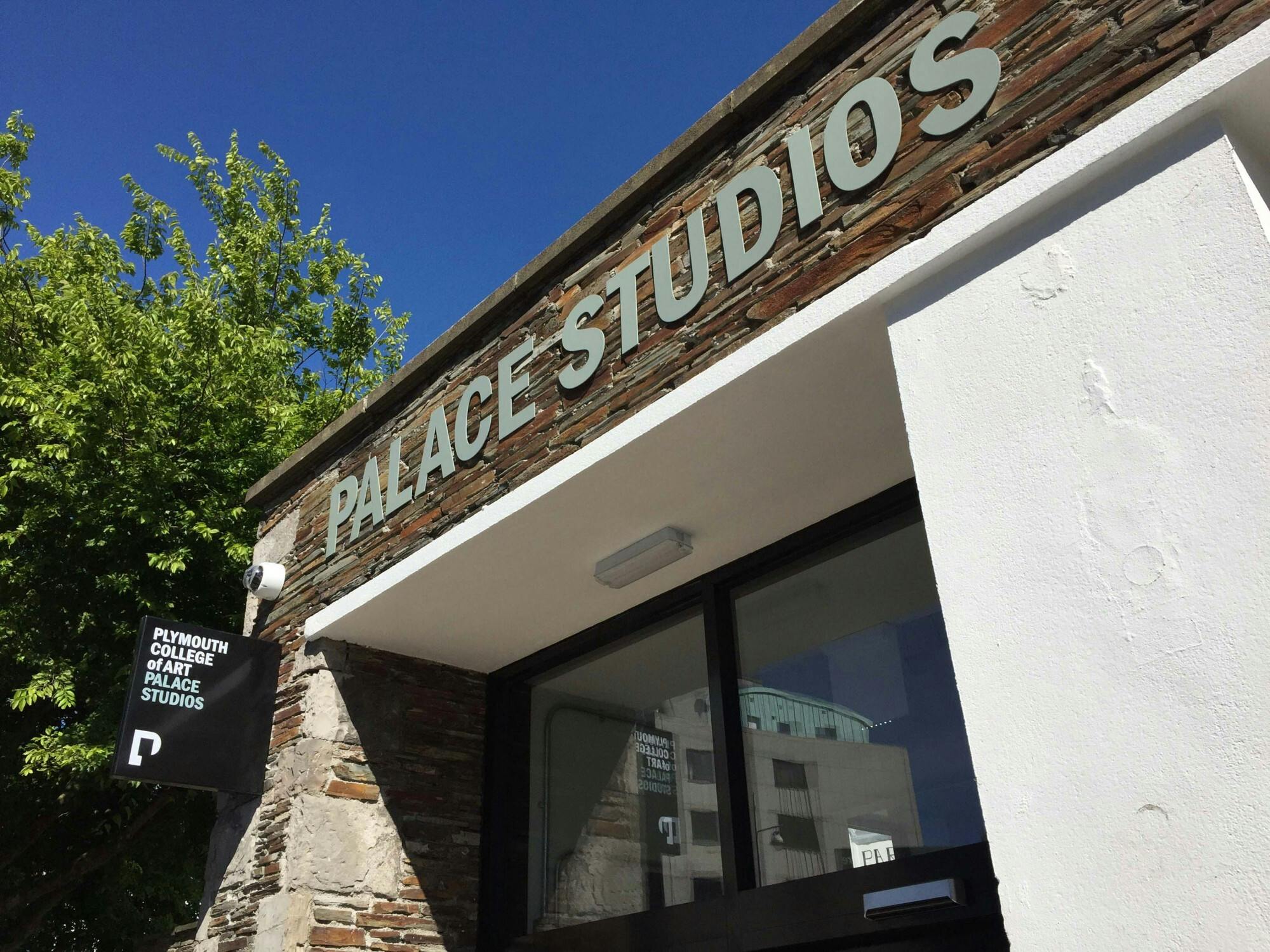 Gazing upwards at the main entrance, a bright blue sky and green leaves frame block caps metal signage that states "Palace Studios" for visitors.