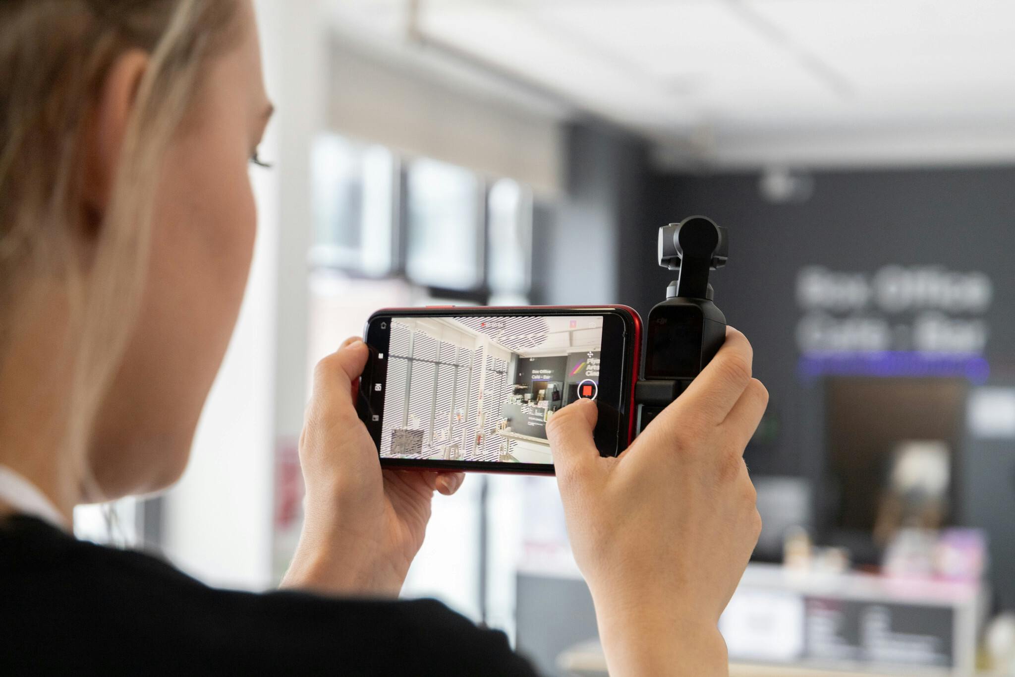 Female student records on an iphone, the device is held landscape in a grip to stabilise hand shakes. The camera screen is sharp and in focus, displaying the entrance way to the college’s on-site cinema.